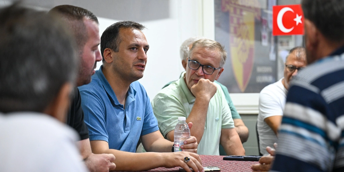 Mavi polo yaka tişört giyen Başkan Eşki, bir masanın etrafında toplanmış esnafla sohbet ederken, yanında gözlüklü bir beyefendi ve diğer katılımcılar dikkatle dinliyor. Arka planda duvarda asılı Türk bayrağı ve bir amblem yer alıyor.