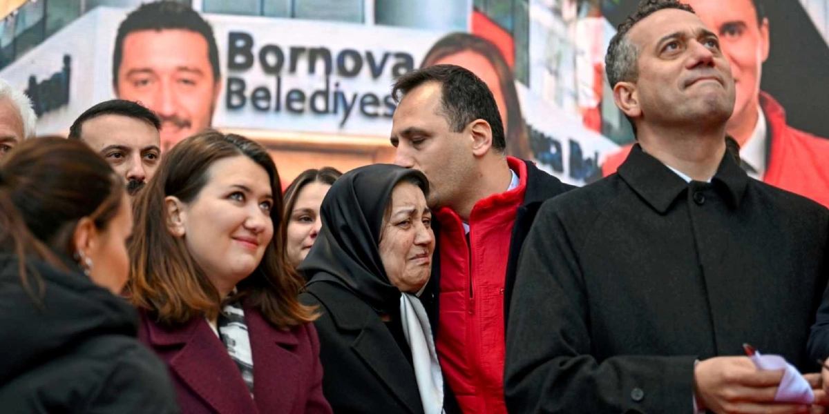 Bornova Belediyesi yazılı bir binanın önünde, kırmızı yelekli bir erkek, başörtülü ve duygulanarak ağlayan bir kadını teselli etmek için başından öpüyor. Yanlarında koyu renkli kaban giymiş, yukarı bakan bir erkek ve bordo ceketli bir kadın yer alıyor. Arka planda Bornova Belediyesi'ne ait görsellerin bulunduğu bir afiş bulunuyor.