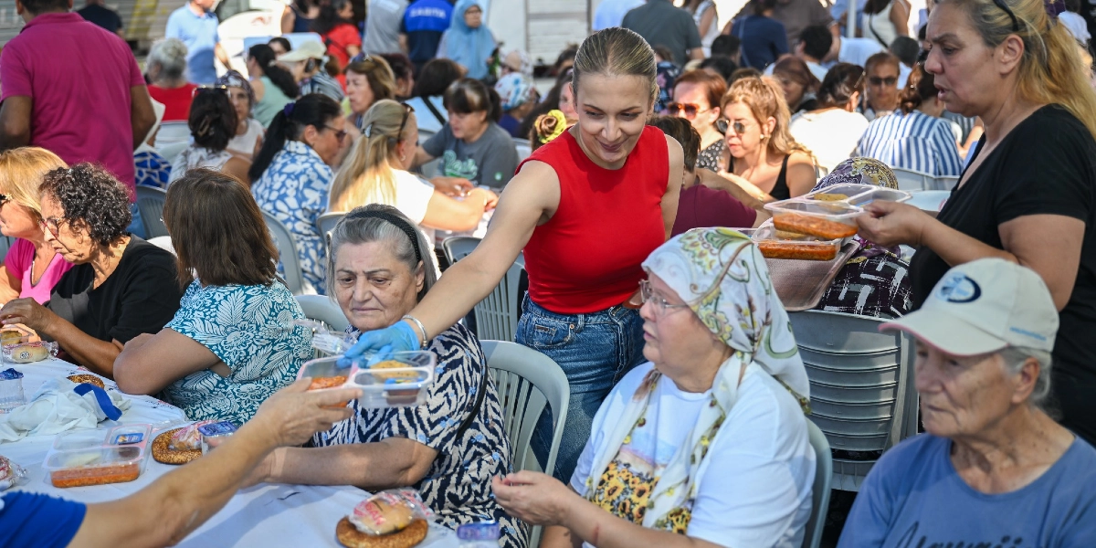 Kırmızı kolsuz bluz giymiş bir kadın, açık havada uzun masalarda oturan kadınlara plastik kaplarda yemek servisi yapıyor. Masalarda oturan kadınlar arasında başörtülü olanlar ve şapka takanlar bulunuyor, önlerinde ise simit ve yemek kapları yer alıyor. Arka planda ise etkinliğe katılan çok sayıda insan görülüyor.