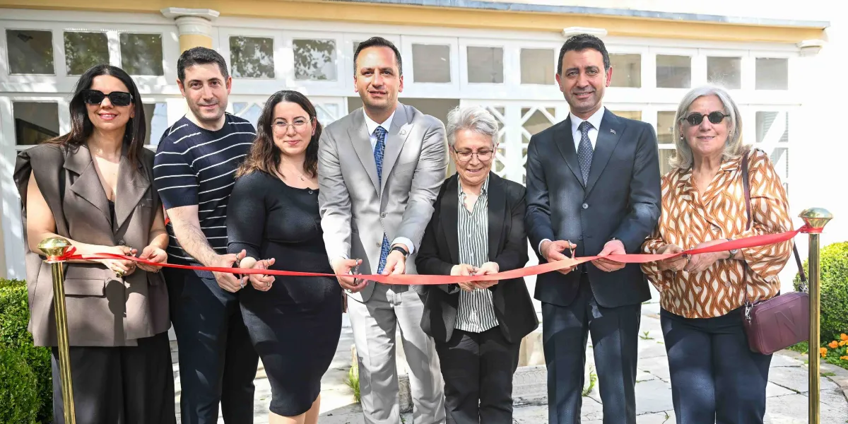 Beyaz bir binanın önünde yan yana duran altı kişilik bir grup, ellerindeki makaslarla kırmızı bir açılış kurdelesini kesiyor. Grupta yer alan üç erkekten ikisi takım elbiseli, biri ise çizgili bir tişört giyiyor. Üç kadın ise günlük şık kıyafetlerle kurdeleyi tutuyor. Hepsi gülümseyerek objektife bakıyor.