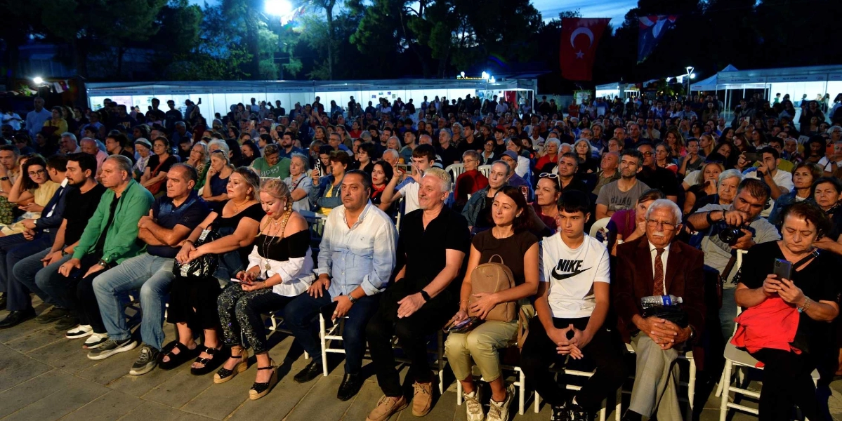 Açık havada, akşam saatlerinde düzenlenen bir etkinlikte, sandalyelerde oturan ve ayakta duran kalabalık bir izleyici grubu sahneye doğru bakıyor. Arka planda beyaz çadırlar ve asılı bir Türk bayrağı görülüyor. İzleyiciler arasında İlber Ortaylı da ön sıralarda, beyaz gömlekli bir beyefendinin yanında oturuyor. Katılımcıların çoğu dikkatle dinliyor, bazıları ise cep telefonlarıyla fotoğraf çekiyor.
