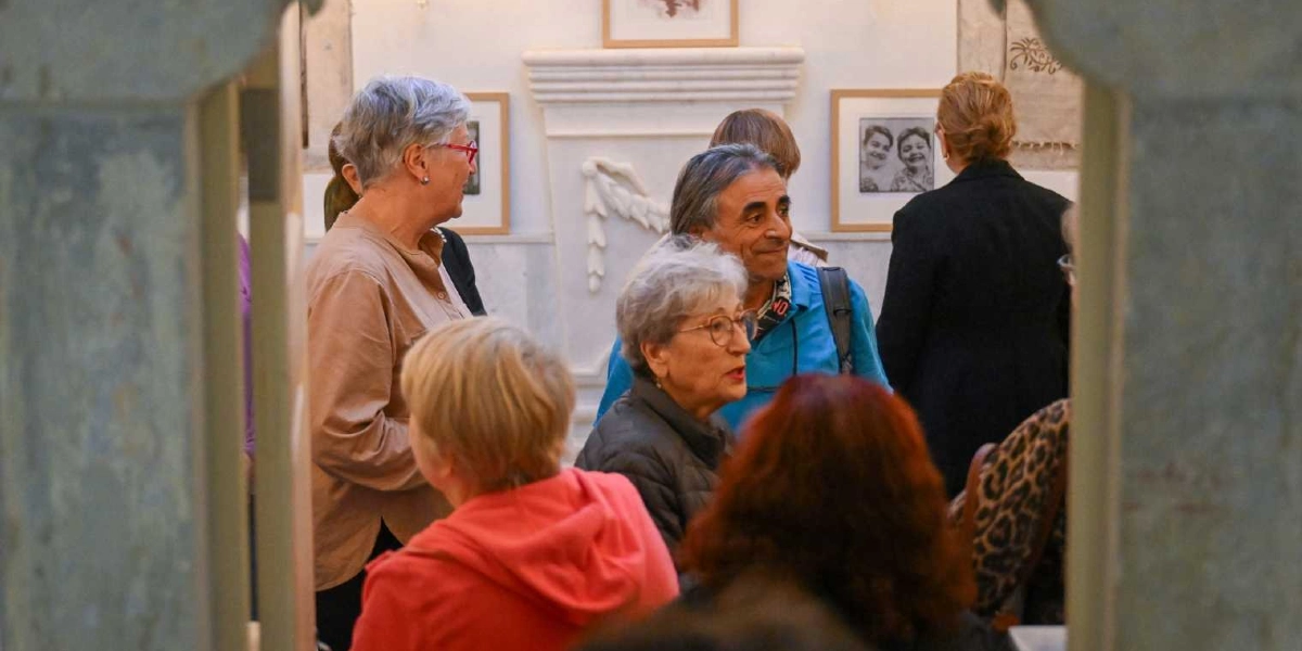Bornova'daki bir sanat galerisinde düzenlenen sergide, bir araya gelen sanatseverler eserleri inceliyor. Ön planda kırmızı kapüşonlu bir kişi ve yanında gri saçlı, gözlüklü bir kadın bulunurken, orta kısımda mavi tişörtlü bir adam ve yanında bej renkli gömlekli, kırmızı gözlüklü bir kadın yer alıyor. Arka planda ise duvarda asılı çerçeveli tablolar ve mermer bir şömine detayı görülüyor.