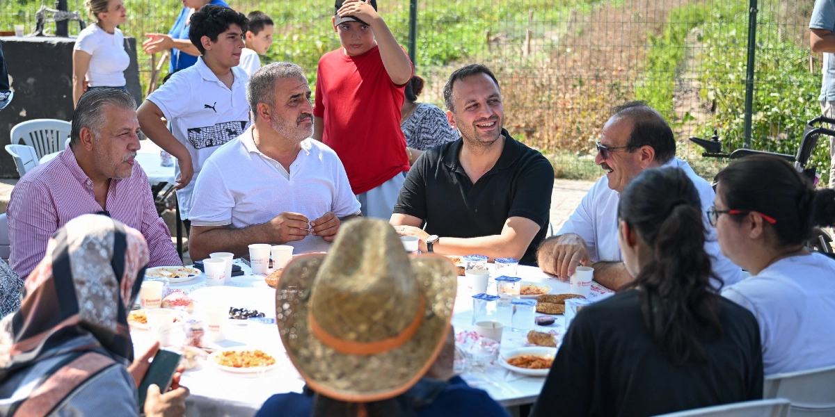 Açık havada, beyaz örtülü uzun bir masanın etrafında toplanmış bir grup insan sohbet ediyor. Masada yemek tabakları, bardaklar ve Bornova Belediyesi logolu su kapları bulunuyor. Ön planda hasır şapkalı bir kişi, orta kısımda ise beyaz gömlekli ve siyah tişörtlü erkekler dikkat çekiyor. Arka planda ise gençlerin ve diğer katılımcıların yer aldığı, tel örgülerle çevrili bir bahçe ortamı görülüyor.