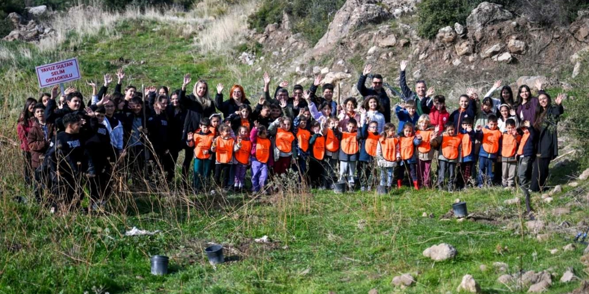 Yavuz Sultan Selim Ortaokulu öğrencileri, öğretmenleri ve yetişkinlerden oluşan kalabalık bir grup, ağaç dikim etkinliği kapsamında açık arazide ellerini havaya kaldırarak poz veriyor. Grubun bir kısmı üzerinde okulun ismi yazılı olan turuncu yelekler giyiyor ve sol tarafta okulun isminin yazılı olduğu bir tabela tutuluyor. Arka planda kayalık ve yeşil bir yamaç bulunuyor.