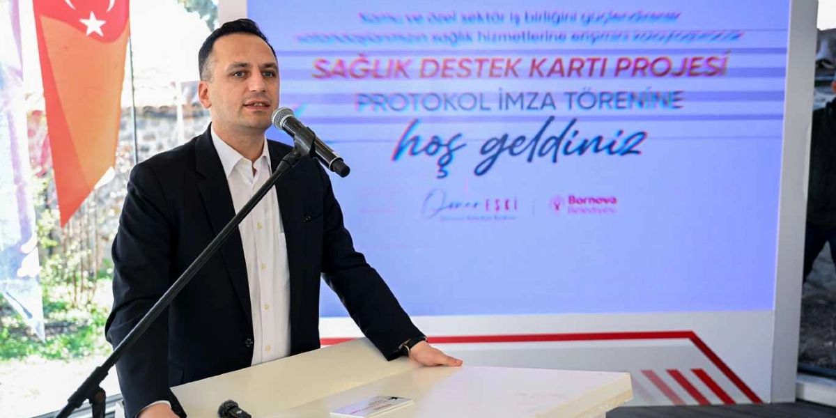 Koyu renk ceket ve beyaz gömlek giymiş bir erkek, kürsüde mikrofon başında konuşma yapıyor. Arkasındaki ekranda 