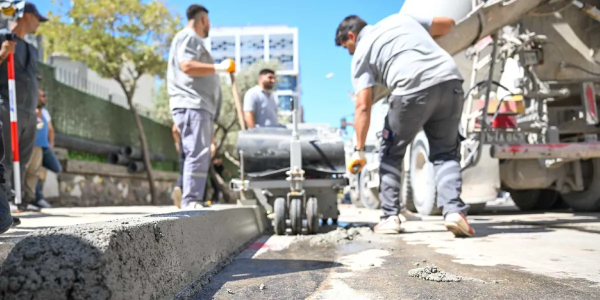 Bornova Belediyesi ekipleri, bir beton dökme makinesi kullanarak yol kenarına yeni bordür taşı döşeme çalışması yapıyor. İşçiler gri üniformalar giymiş durumda ve yanlarında beton mikseri aracı bulunuyor.