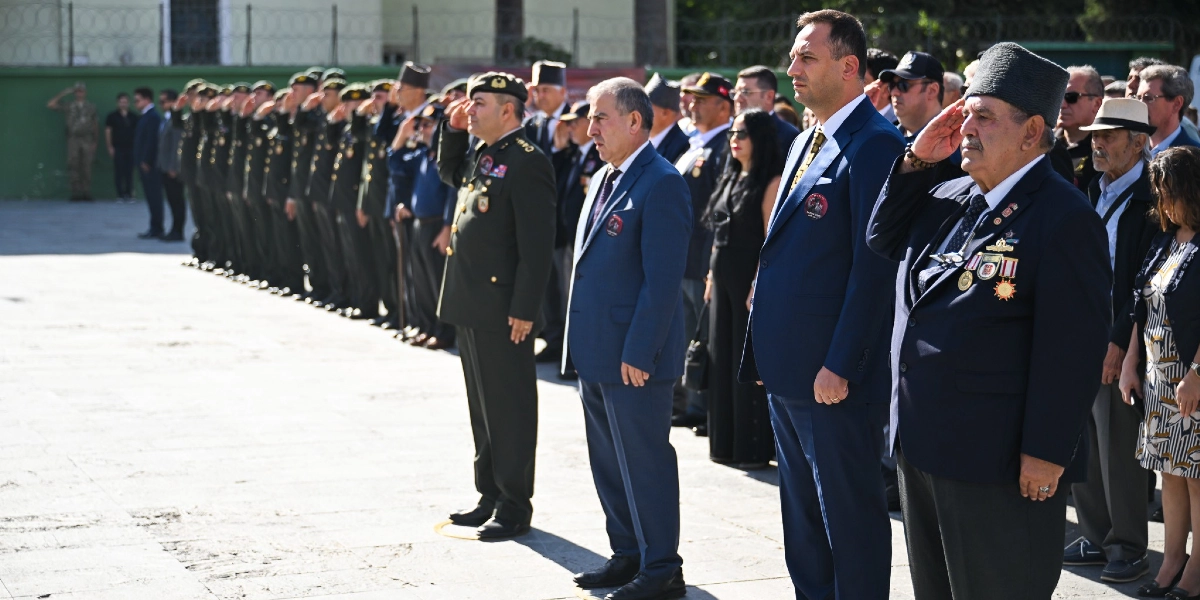 Açık havada düzenlenen 19 Eylül Gaziler Günü töreninde, askeri üniformalı askerler ve sivil kıyafetli gaziler yan yana dizilmiş şekilde selam duruyor. Ön planda, üzerinde madalyalar bulunan koyu renk takım elbiseli ve kalpaklı bir gazi, yanında lacivert takım elbiseli yetkililer ve askeri üniformalı bir subay ile birlikte saygı duruşunda bulunuyor. Arka planda ise resmi tören düzeninde sıralanmış askerler yer alıyor.