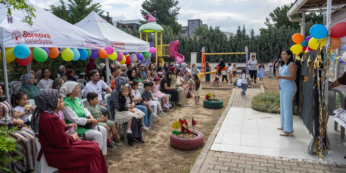 Bornova Belediyesi logolu beyaz çadırların altında, sandalyelerde oturan aileler ve çocuklar bir etkinlik için toplanmış. Açık hava alanında, elinde mikrofonla konuşma yapan açık mavi takım elbiseli bir kadın bulunuyor. Arka planda çocukların oynadığı bir park alanı ve renkli balonlarla süslenmiş bir ortam görülüyor.