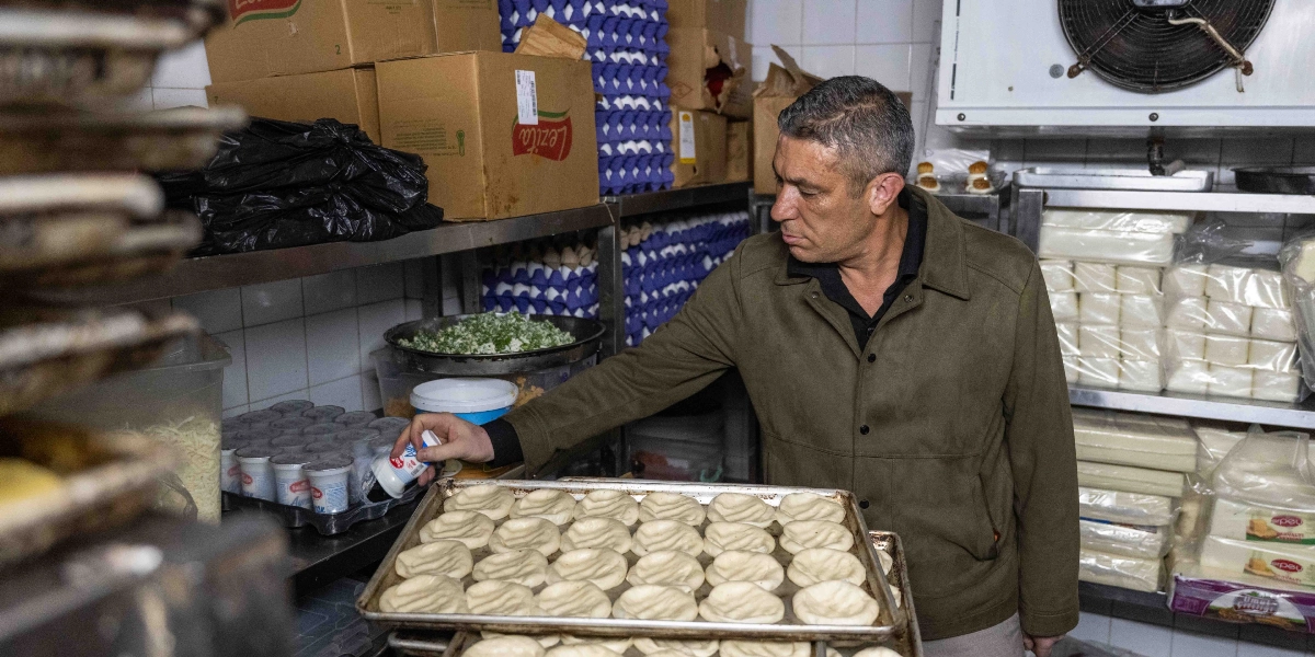 Kısa saçlı, zeytin yeşili ceket giymiş bir adam, mutfak tezgahında dizili hamur parçalarının yanındaki küçük bir yoğurt kabını eline alıyor. Arka planda raflarda dizili yumurta kolileri, peynir paketleri ve çeşitli gıda malzemeleri bulunuyor.