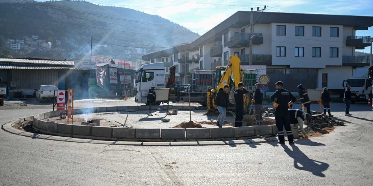 Bornova'da bir yol kavşağında devam eden düzenleme çalışmaları sırasında, iş kıyafetli bir grup işçi dairesel bir refüj çevresinde çalışıyor. Arka planda sarı renkli bir iş makinesi, park halindeki tırlar ve yeni inşa edilmiş apartmanlar yer alıyor. Çalışma alanının etrafında trafik levhaları ve duman çıkaran küçük bir ateş görülüyor.