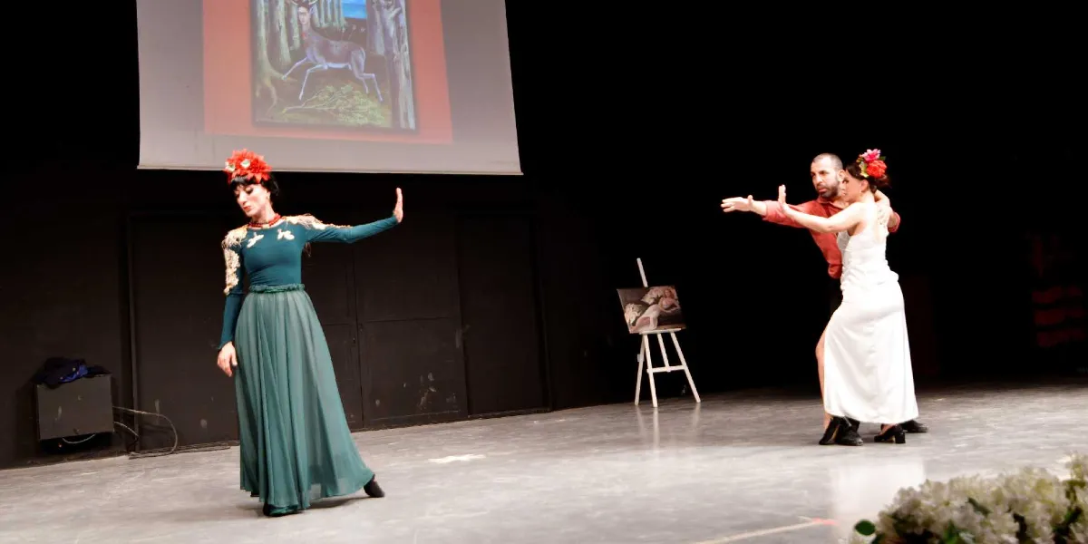 Karanlık bir sahnede, Frida Kahlo'yu andıran saç aksesuarlı ve yeşil uzun elbiseli bir kadın dansçı ile beyaz elbiseli bir kadın ve partneri olan erkek dansçının sergilediği performans. Arka planda Frida Kahlo'nun 