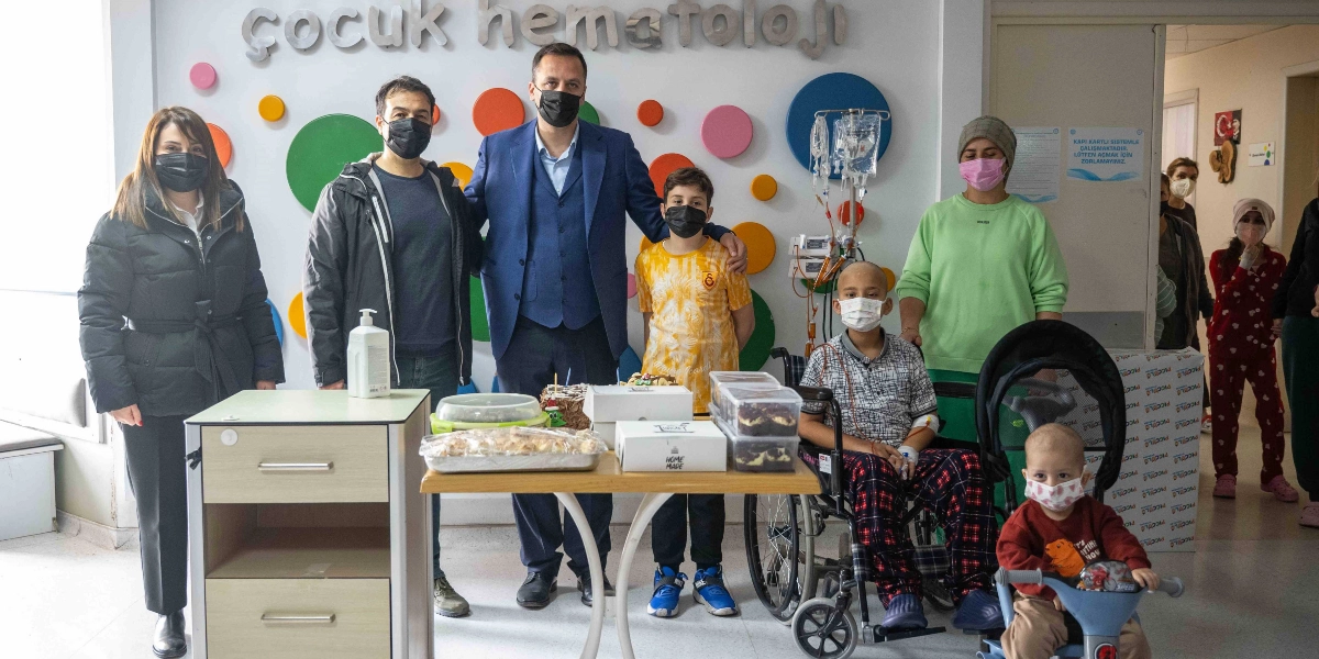 Bornova Çocuk Hematoloji servisinde, üzerinde çeşitli yiyeceklerin bulunduğu bir masanın etrafında toplanmış bir grup yetişkin ve çocuk. Arka planda duvarda renkli daireler ve 
