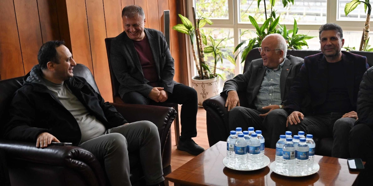 Ahşap kaplamalı bir ofis ortamında, deri koltuklarda oturan dört erkek samimi bir sohbet gerçekleştiriyor. Önlerindeki alçak masada iki tepsi üzerinde çok sayıda küçük plastik su şişesi bulunuyor. Arka planda büyük pencerelerin önünde saksıda yeşil bitkiler yer alıyor.