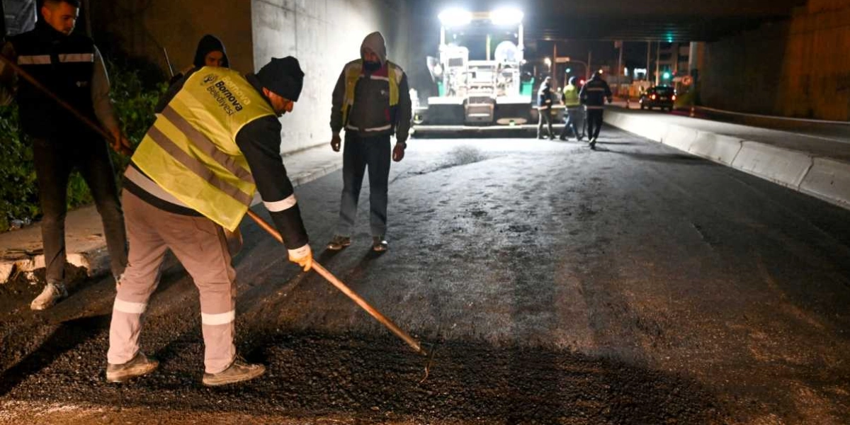 Gece saatlerinde bir alt geçitte çalışan Bornova Belediyesi ekipleri, ellerindeki tırmıklarla yeni serilen asfaltı düzeltiyor. İşçiler reflektörlü yelekler giyiyor ve arka planda asfalt serme makinesi ile diğer görevliler görülüyor.