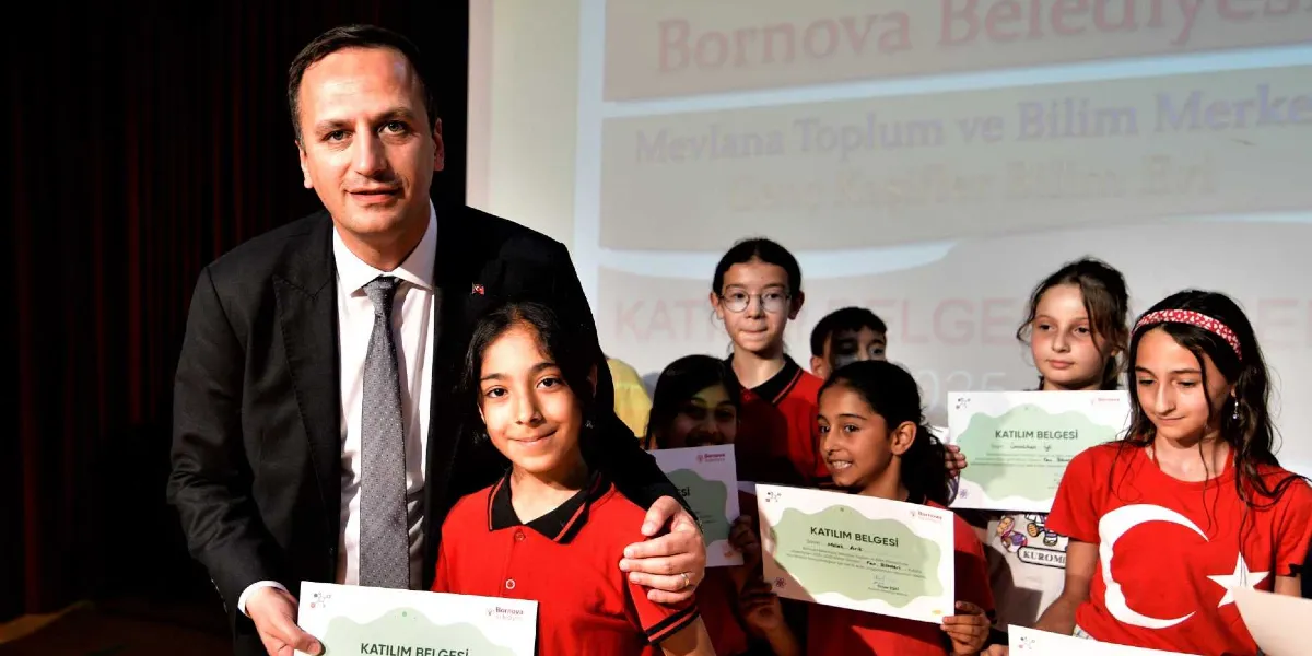 Bornova Belediye Başkanı, Mevlana Toplum ve Bilim Merkezi'nde düzenlenen törende, ellerinde katılım belgeleri bulunan çocuklara sertifikalarını takdim ediyor. Arka planda Bornova Belediyesi ve Mevlana Toplum ve Bilim Merkezi yazılı bir projeksiyon ekranı yer alıyor. Çocuklar kırmızı tişörtler giyiyor ve içlerinden biri Türk bayraklı bir tişörtle dikkat çekiyor.