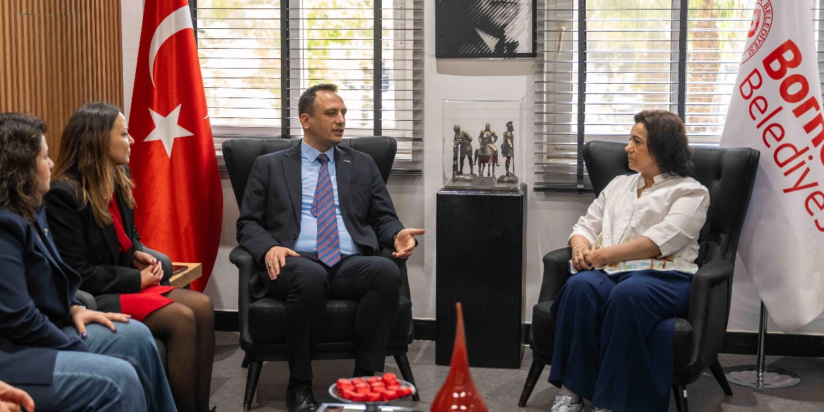 Ofis ortamında bir araya gelen dört kişi, siyah koltuklarda oturarak sohbet ediyor. Ortada lacivert takım elbiseli ve çizgili kravatlı bir erkek, karşısında beyaz bluz ve lacivert pantolon giyen bir kadın ile yan yana oturan iki kadın yer alıyor. Arka planda Türk bayrağı ve üzerinde Bornova Belediyesi yazılı bir flama bulunuyor. İkili arasında, cam bir fanus içerisinde asker figürlerinden oluşan bir heykel sergileniyor.