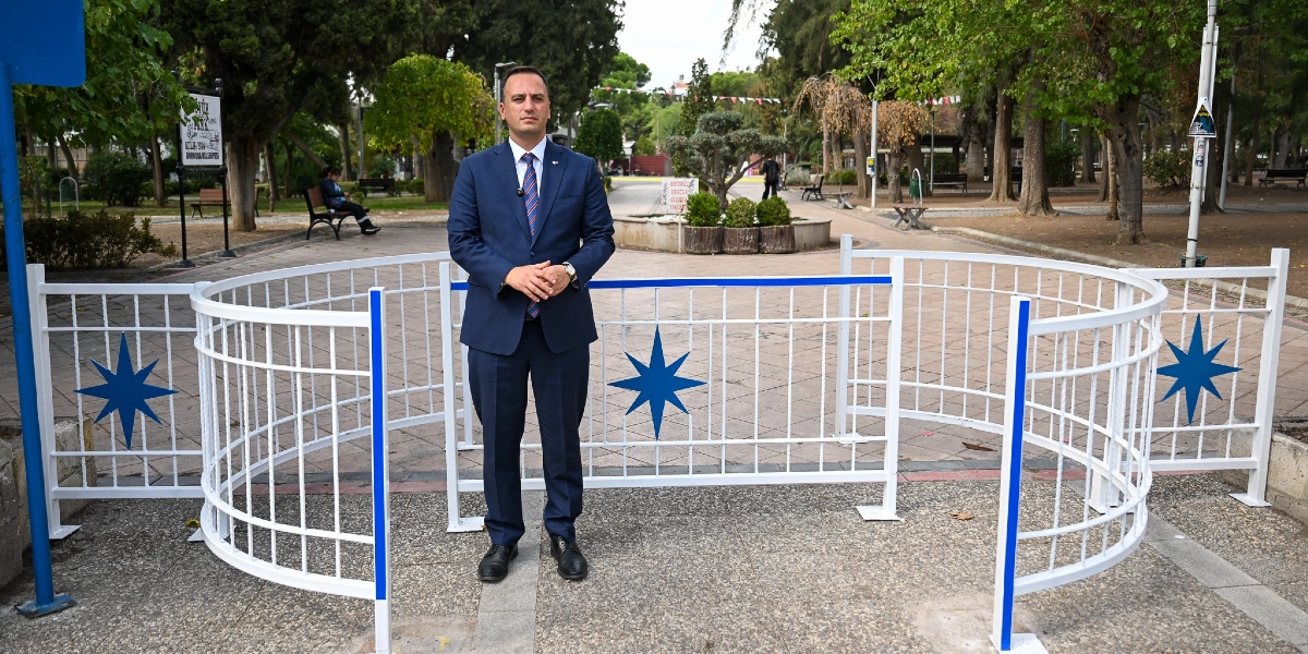 Lacivert takım elbiseli, kravatlı bir erkek, Bornova'da bir parkın girişine yerleştirilmiş, üzerinde mavi yıldız motifleri bulunan beyaz renkli yeni güvenlik bariyerlerinin önünde duruyor. Bariyerler, motosikletlerin parka girişini engellemek amacıyla zikzaklı bir geçiş düzeni oluşturacak şekilde konumlandırılmış.
