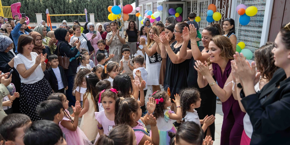 Bir anaokulu veya kreş bahçesinde, renkli balonlarla süslenmiş bir ortamda, öğretmenler ve veliler çocukların karne veya belge alma sevincine alkışlarla eşlik ediyor. Ön planda şenlikli kıyafetler içindeki çocuklar ve onları neşeyle izleyen yetişkinler yer alıyor. Bazı veliler anı ölümsüzleştirmek için telefonlarıyla fotoğraf çekiyor, ortamda genel bir kutlama havası hakim.