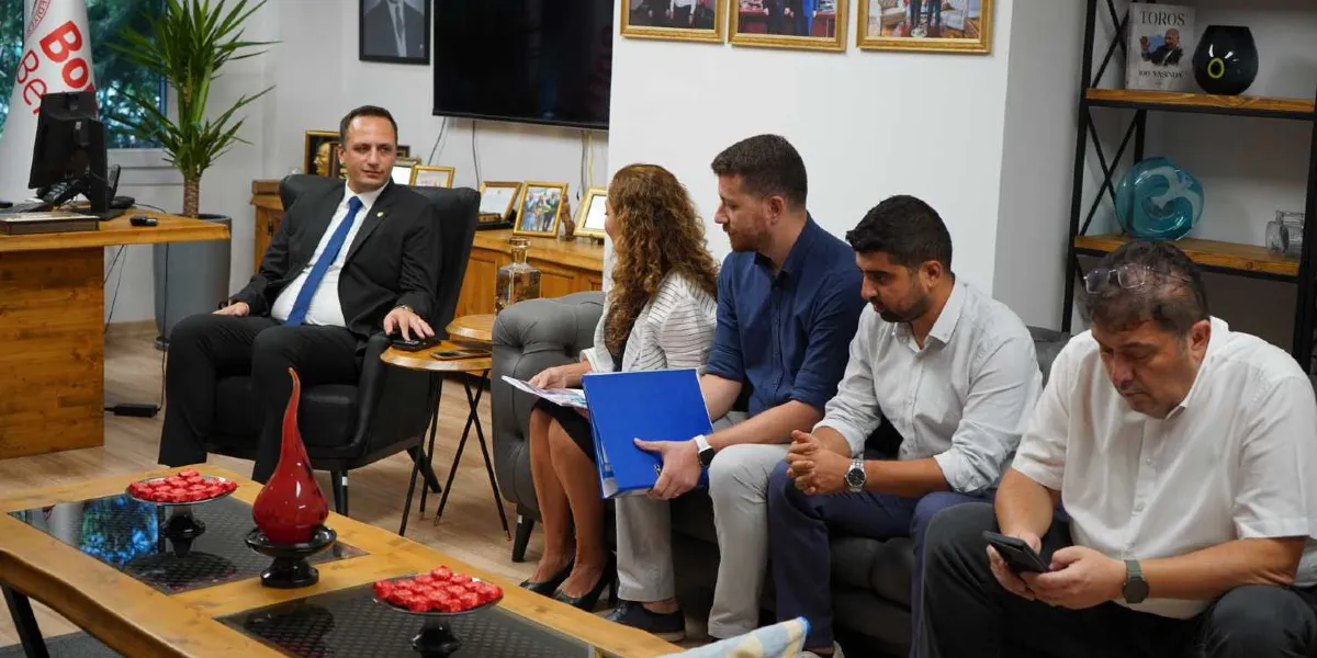 Ofis ortamında bir toplantı sırasında, takım elbiseli bir yönetici tekli koltukta otururken, karşısındaki gri renkli kanepede dört kişilik bir grup yer alıyor. Kanepede oturanlardan biri elinde mavi bir dosya tutarken, diğerleri dikkatle dinliyor veya telefonlarıyla ilgileniyor. Önlerindeki ahşap sehpada kırmızı süslemeli tabaklar ve dekoratif bir obje bulunuyor. Arka planda duvarda çerçeveli fotoğraflar, kitaplıkta çeşitli aksesuarlar ve bir televizyon ekranı yer alıyor.