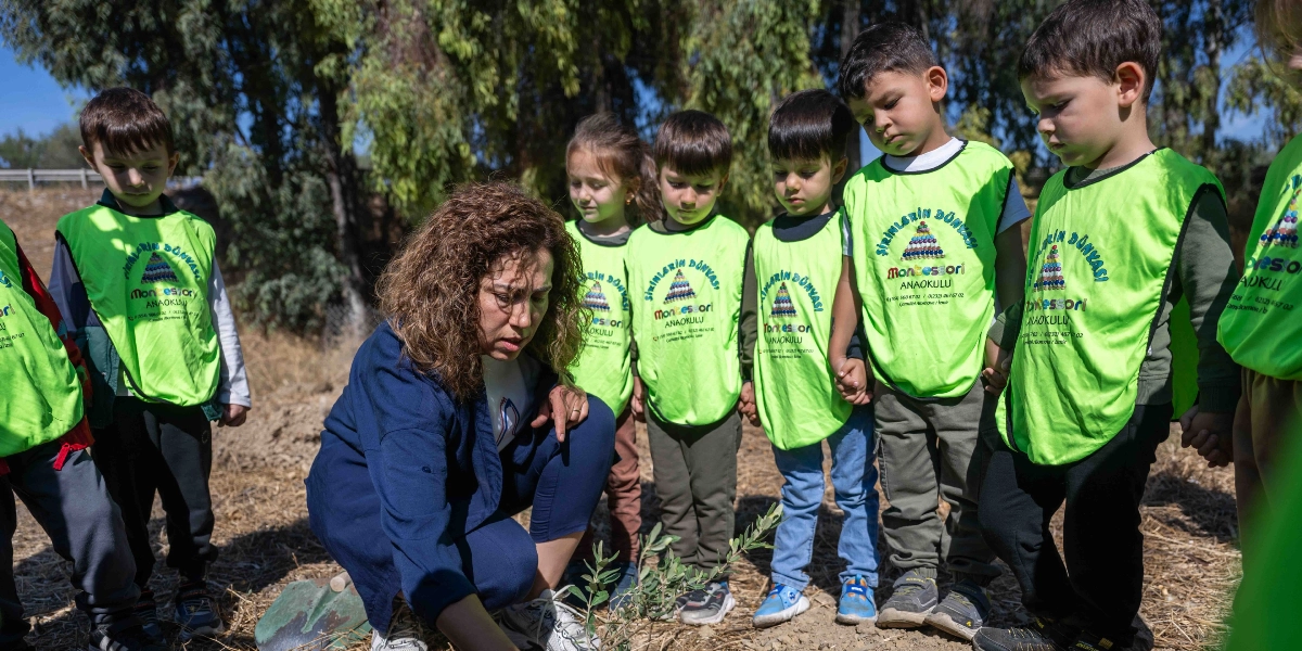 Üzerinde Şirinlerin Dünyası Montessori Anaokulu yazılı neon yeşil yelekler giymiş bir grup çocuk, ağaçlık bir alanda yere çömelmiş bir öğretmeni izliyor. Öğretmen, toprağa yeni dikilmiş küçük bir fidanla ilgilenirken, çocuklar meraklı bakışlarla onu takip ediyor.