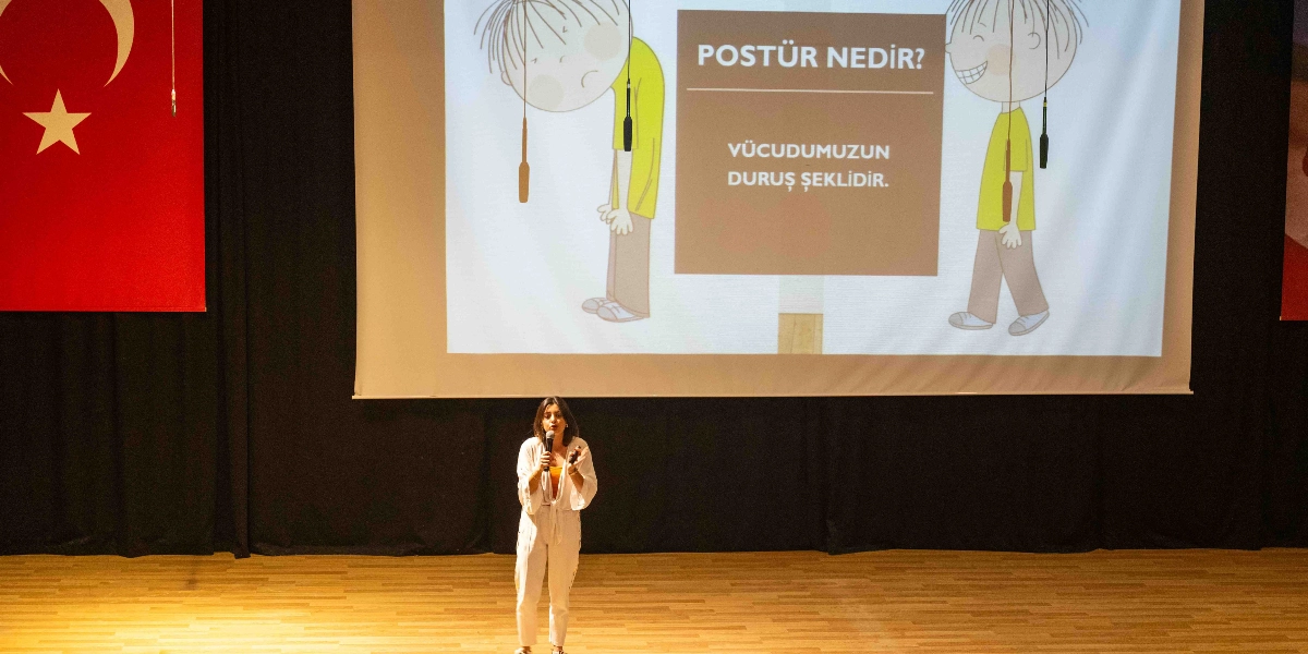 Sahne üzerinde elinde mikrofonla sunum yapan genç bir kadın ve arkasındaki dev ekranda postürün tanımını yapan eğitici bir görsel yer alıyor. Ekranda, biri kambur duran diğeri ise dik duran iki çocuk çizimi ile 
