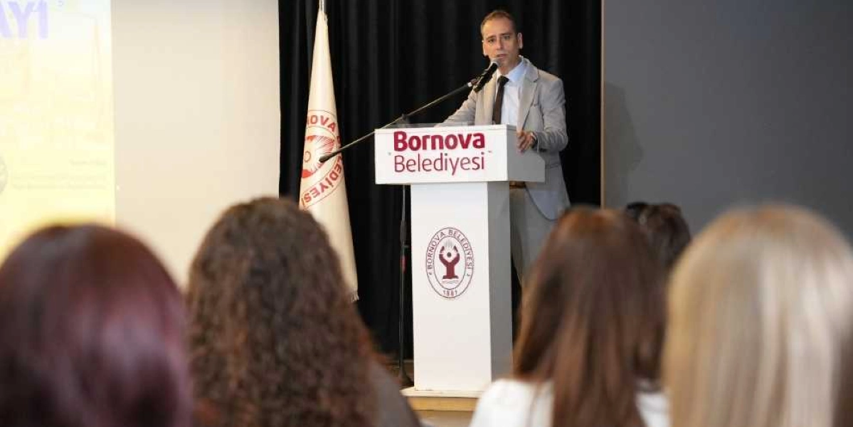 Gri takım elbiseli ve kravatlı bir konuşmacı, üzerinde Bornova Belediyesi yazan ve belediyenin logosunu taşıyan bir kürsüde, dinleyici kitlesine hitap ediyor. Kürsünün hemen yanında Bornova Belediyesi bayrağı yer alıyor. Ön planda ise dinleyicilerin başlarının arkası bulanık bir şekilde görülüyor.