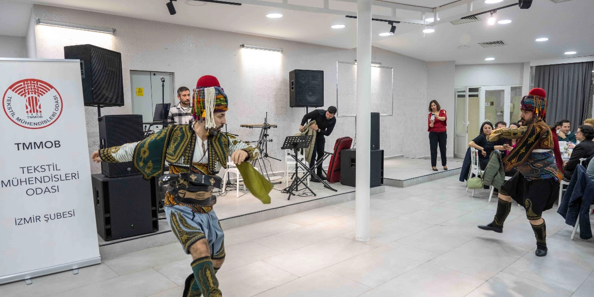 TMMOB Tekstil Mühendisleri Odası İzmir Şubesi'nin düzenlediği bir etkinlikte, geleneksel kıyafetler giymiş iki erkek halk oyunu sergiliyor. Salonun sol tarafında üzerinde kurumun logosu ve ismi bulunan bir tanıtım panosu yer alıyor. Arka planda müzik ekipmanları ve hazırlık yapan görevliler bulunurken, sağ taraftaki masalarda oturan davetliler gösteriyi izliyor.