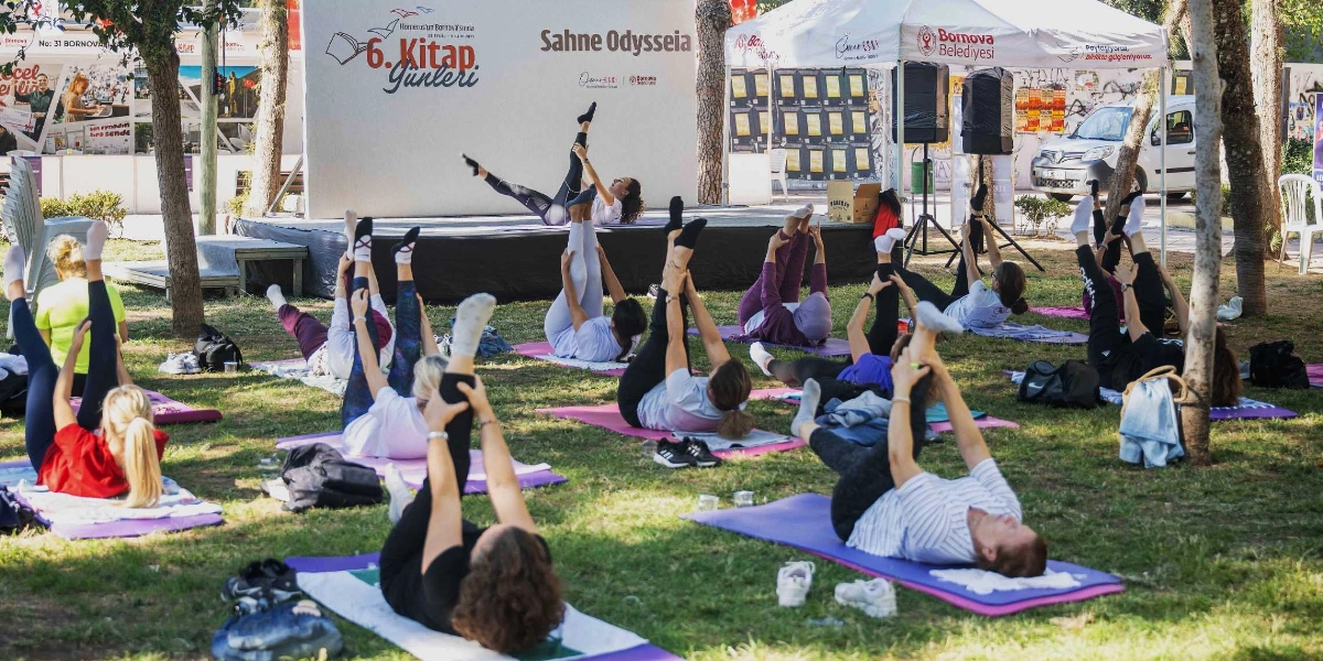 Bornova Belediyesi tarafından düzenlenen 6. Kitap Günleri kapsamında, ağaçlık bir alanda yoga matları üzerinde egzersiz yapan bir grup insan. Arka planda 