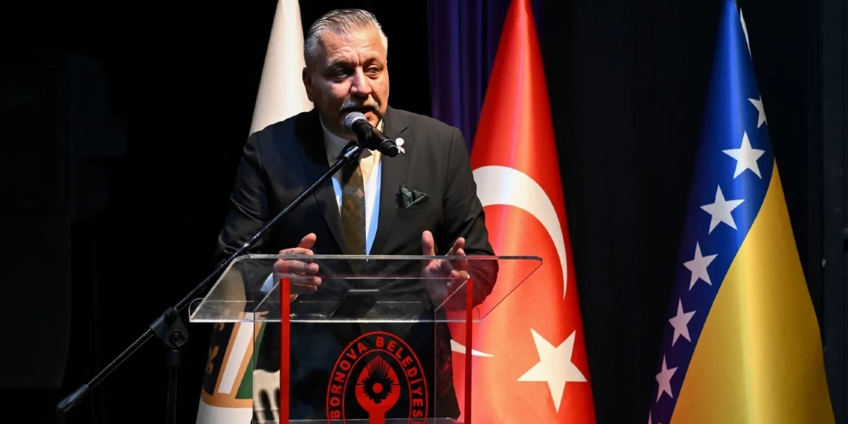 Koyu renkli takım elbise ve kravat giymiş, bıyıklı bir erkek, üzerinde Bornova Belediyesi logosu bulunan şeffaf bir kürsüde mikrofon başında konuşma yapıyor. Arka planda Türkiye Cumhuriyeti bayrağı ile Bosna Hersek bayrağı yer alıyor.