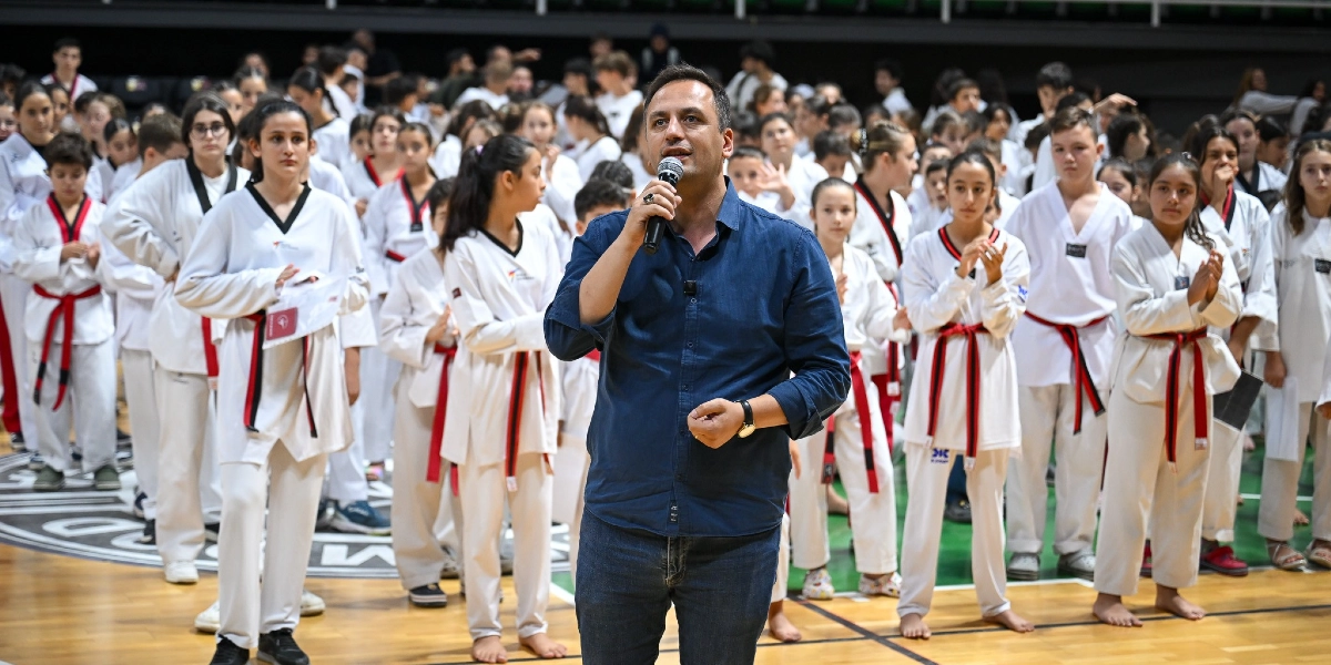 Kapalı bir spor salonunda, lacivert gömlekli bir adam elinde mikrofonla konuşma yaparken, arkasında tekvando kıyafetleri ve kırmızı-siyah kuşakları giymiş kalabalık bir sporcu grubu onu dinliyor.