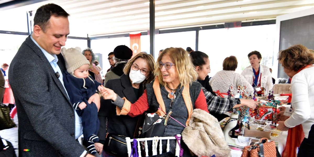 Koyu renk ceketli bir adamın kucağında bebekle gülümsediği, çevresinde ise el yapımı takıların sergilendiği bir stantta sohbet eden kadınların bulunduğu kapalı bir etkinlik alanı. Adamın yanındaki maskeli bir kadın bebeğin elini tutarken, yanındaki diğer kadın onlara gülümseyerek eşlik ediyor. Arka planda Türk bayrağı ve çeşitli eşyaların sergilendiği masalarla ilgilenen başka katılımcılar görülüyor.
