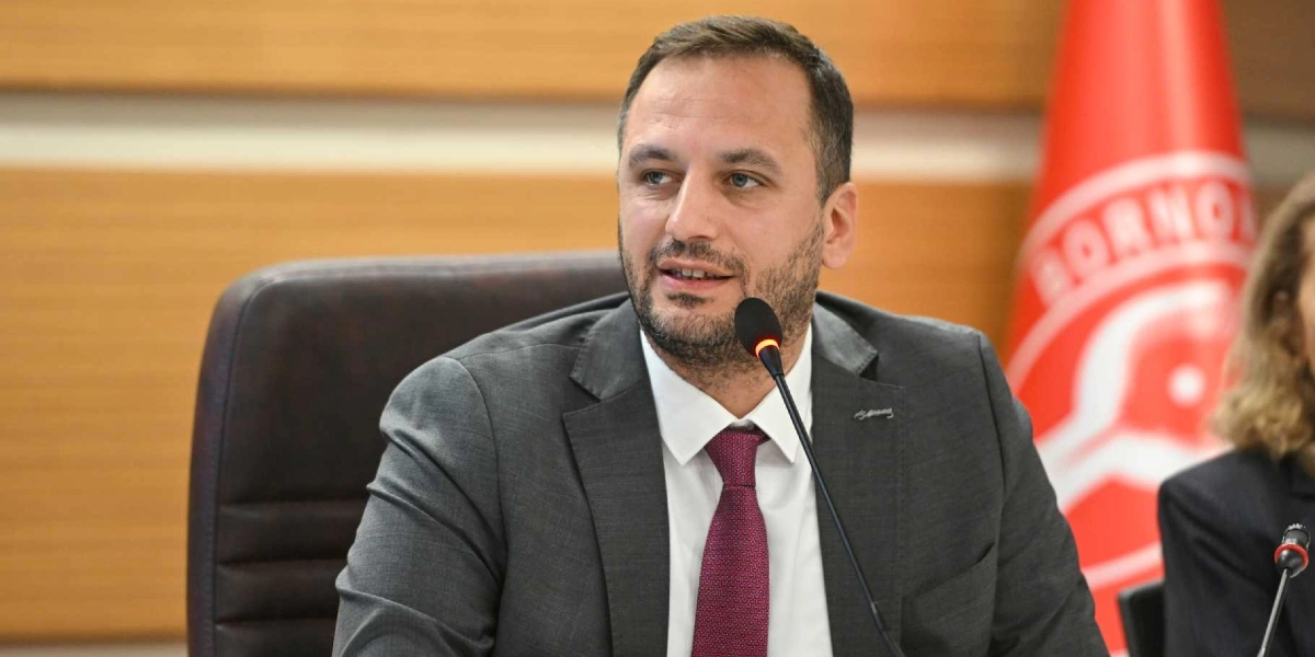 Koyu gri takım elbiseli, beyaz gömlekli ve bordo kravatlı bir erkek, önünde mikrofon bulunan bir masada oturarak konuşma yapıyor. Arka planda Bornova Belediyesi'nin kırmızı renkli logosunun yer aldığı bir bayrak bulunuyor.