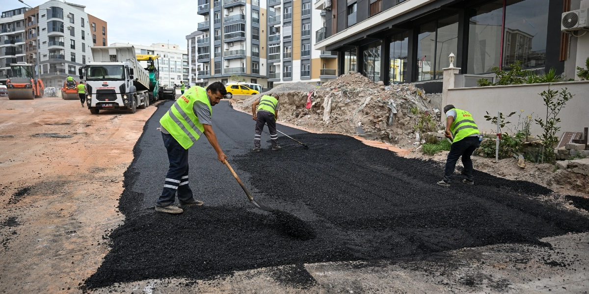 Şehir merkezinde, üzerinde sarı reflektörlü iş yelekleri bulunan üç işçi, yeni dökülen asfaltı kürek ve tırmık yardımıyla düzeltiyor. Arka planda modern apartmanlar, park halindeki araçlar ve asfalt serme çalışmalarında kullanılan bir kamyon ile silindir makinesi görülüyor.