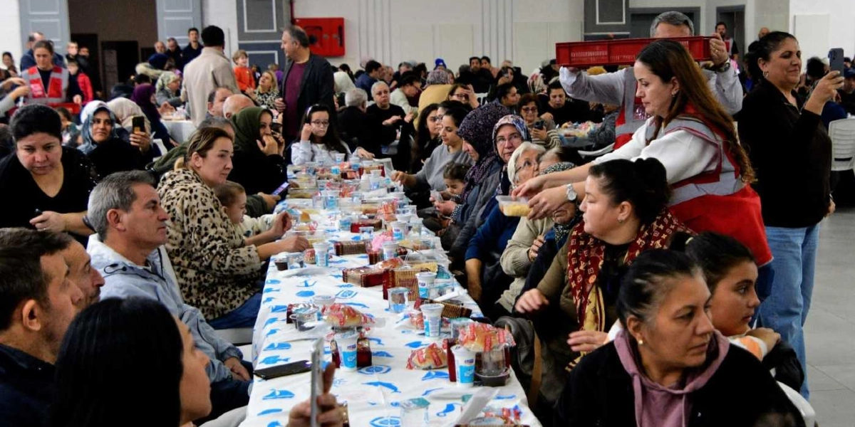Kapalı bir spor salonu veya büyük bir etkinlik alanında, uzun bir masanın etrafında toplanmış kalabalık bir grup insan yılbaşı yemeğinde bir arada oturuyor. Masanın üzerinde paketli yiyecekler, içecekler ve plastik kaplar bulunuyor. Kırmızı yelekli bir görevli, masadaki katılımcılara yemek servisi yapıyor. Katılımcıların bir kısmı yemek yerken, bazıları ise cep telefonlarıyla fotoğraf çekiyor veya sohbet ediyor. Ortamda farklı yaş gruplarından kadınlar, erkekler ve çocuklar yer alıyor.