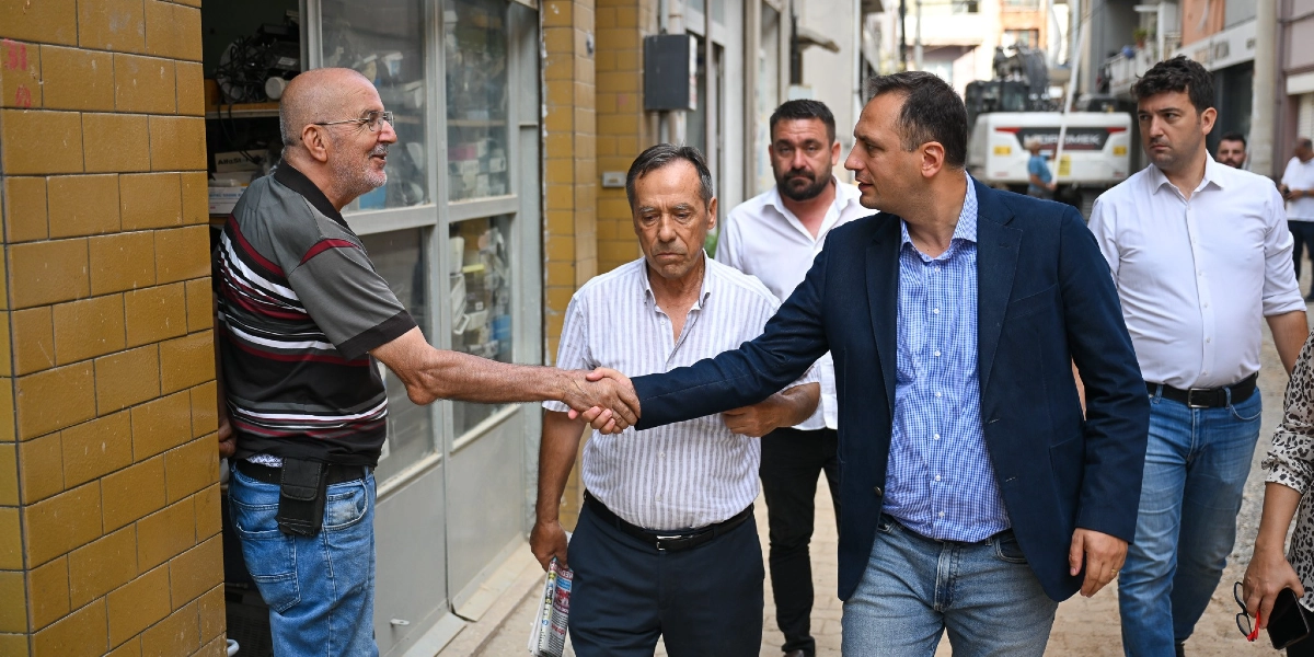 Bornova Belediye Başkanı Mustafa İduğ, bir sokak gezisi sırasında esnafla tokalaşırken görülüyor. Başkan İduğ, mavi ekose gömlek ve lacivert ceket giymiş durumda; yanında ise belediye yetkilileri ve arkada çalışma yapan bir iş makinesi yer alıyor.
