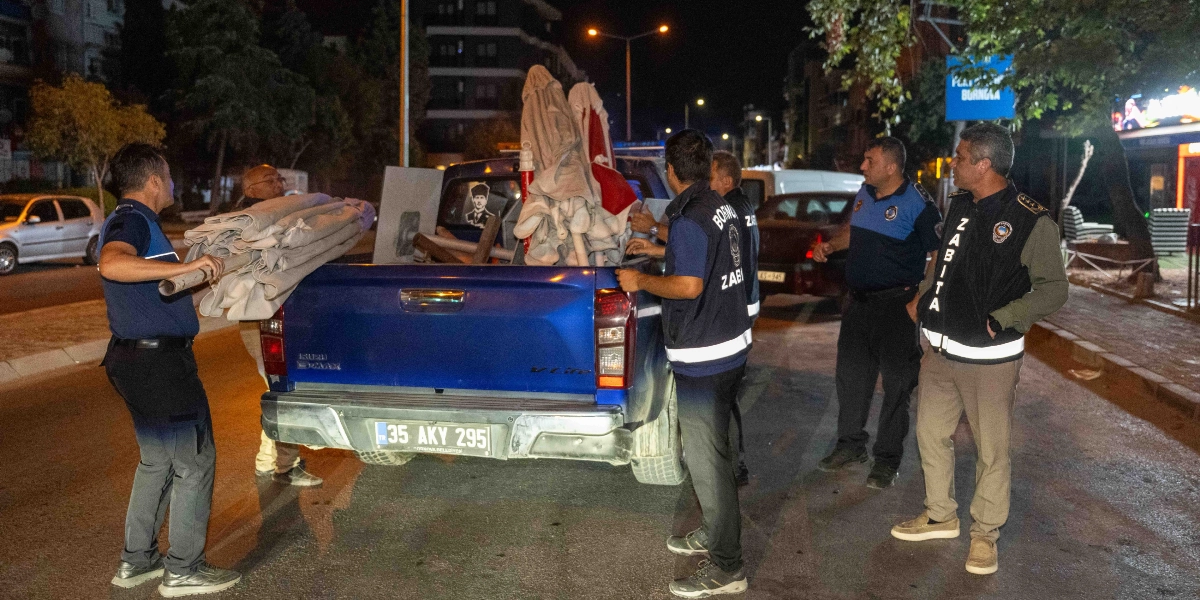 Gece vakti Bornova Belediyesi zabıta ekipleri, kaldırımdan topladıkları şemsiye ve çeşitli malzemeleri 35 AKY 295 plakalı mavi bir kamyonetin kasasına yüklüyor. Üniformalı zabıta memurları operasyon sırasında çevrede bulunuyor.