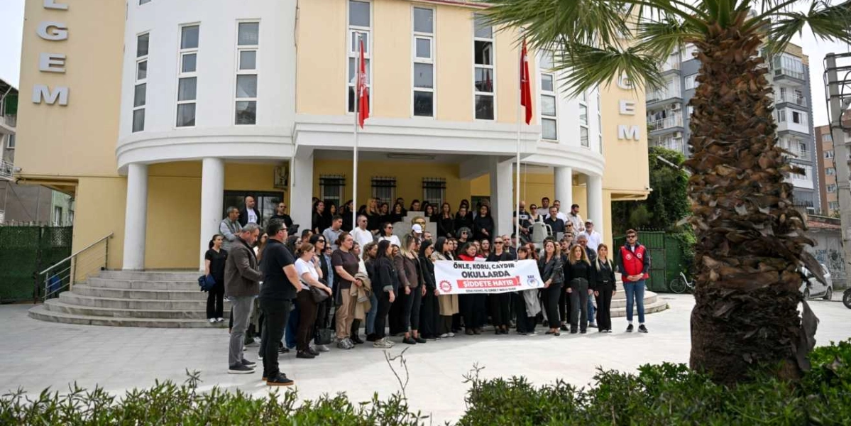 Açık renkli, çok katlı bir okul binasının önünde toplanmış, çoğunluğu siyah kıyafetler giymiş kalabalık bir grup insan, ellerinde 