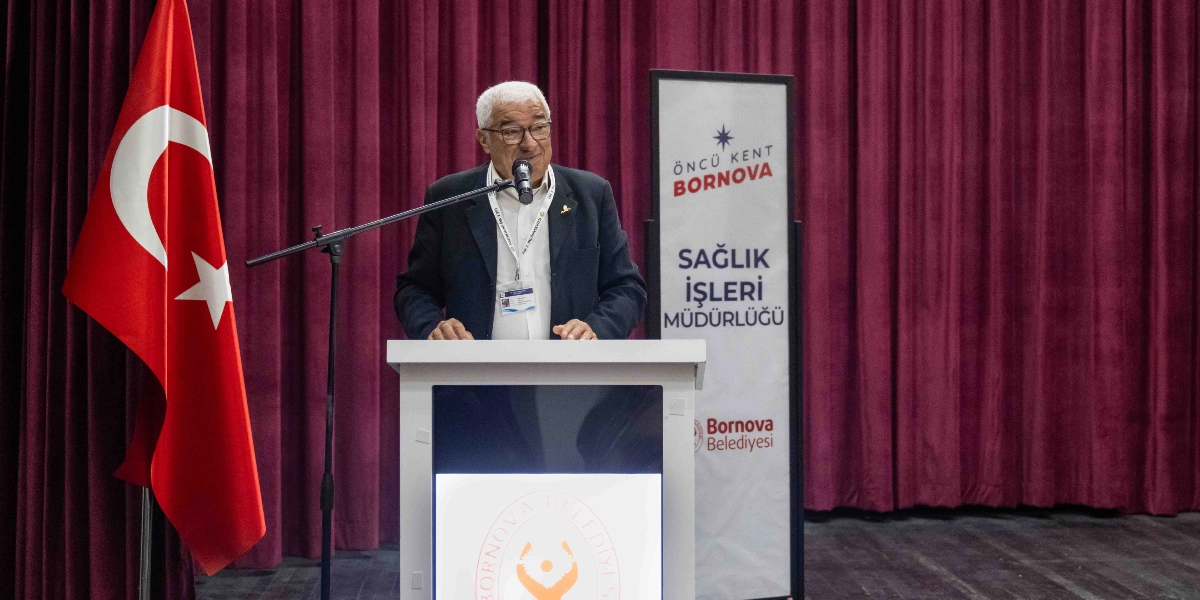 Beyaz saçlı, gözlüklü ve koyu renk ceketli bir konuşmacı, bordo renkli perde fonu önünde yer alan kürsüde mikrofon başında hitap ediyor. Kürsünün solunda Türk bayrağı, sağında ise üzerinde 