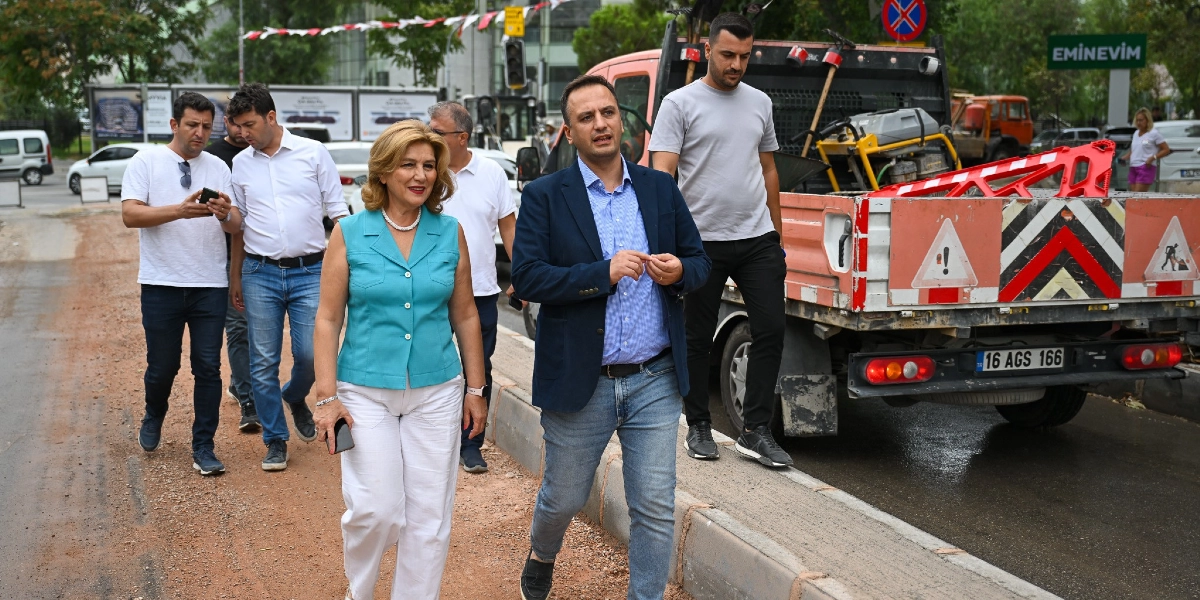Bornova Zafer Caddesi'ndeki düzenleme çalışmalarını inceleyen bir grup yetkili, yol kenarındaki toprak zeminde yürürken görülüyor. Grubun önünde turkuaz kolsuz bir bluz ve beyaz pantolon giyen bir kadın ile mavi gömlek ve lacivert ceket giyen bir erkek yer alıyor. Arkalarında beyaz tişörtlü ve kot pantolonlu iki erkek daha bulunuyor. Yanlarında ise üzerinde inşaat ekipmanları ve uyarı levhaları bulunan, 16 AGS 166 plakalı bir kamyonet park halinde bekliyor.