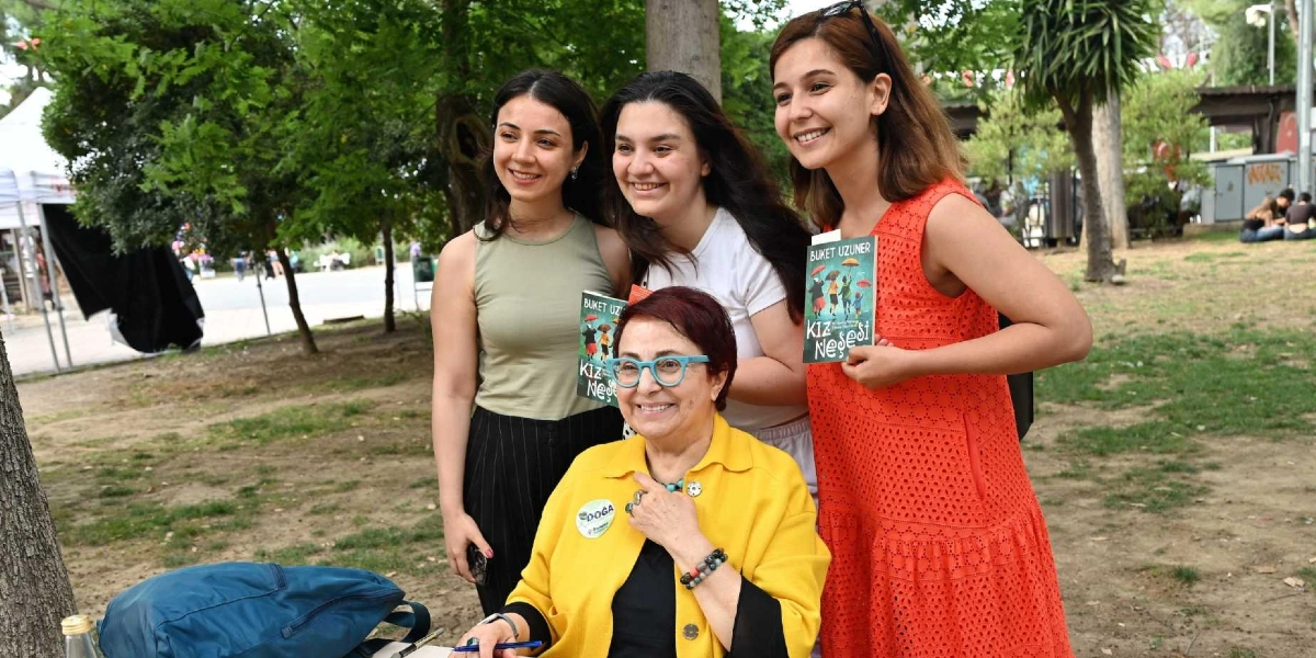 Açık havada, ağaçlık bir alanda yazar Buket Uzuner, üzerinde sarı bir ceketle otururken yanında üç genç kadınla birlikte gülümsüyor. Kadınlardan ikisi ellerinde Buket Uzuner'in 