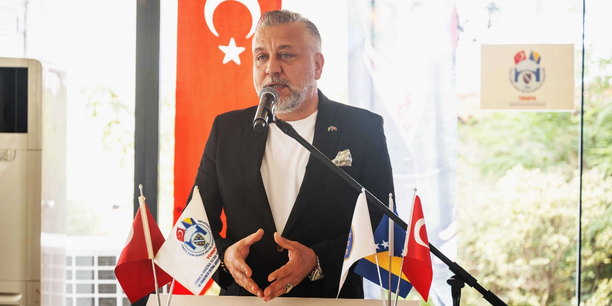 Siyah ceket ve beyaz tişört giymiş orta yaşlı bir erkek, kürsüde mikrofon başında konuşma yapıyor. Arka planda Türk bayrağı ve üzerinde Türkiye ile Bosna Hersek bayraklarının yer aldığı bir kurum logosu bulunuyor. Kürsü üzerinde ise Türkiye ve Bosna Hersek bayrakları ile Türkiye Bosna Hersek Kültür Dernekleri Federasyonu'nun logolu flamaları yer alıyor.