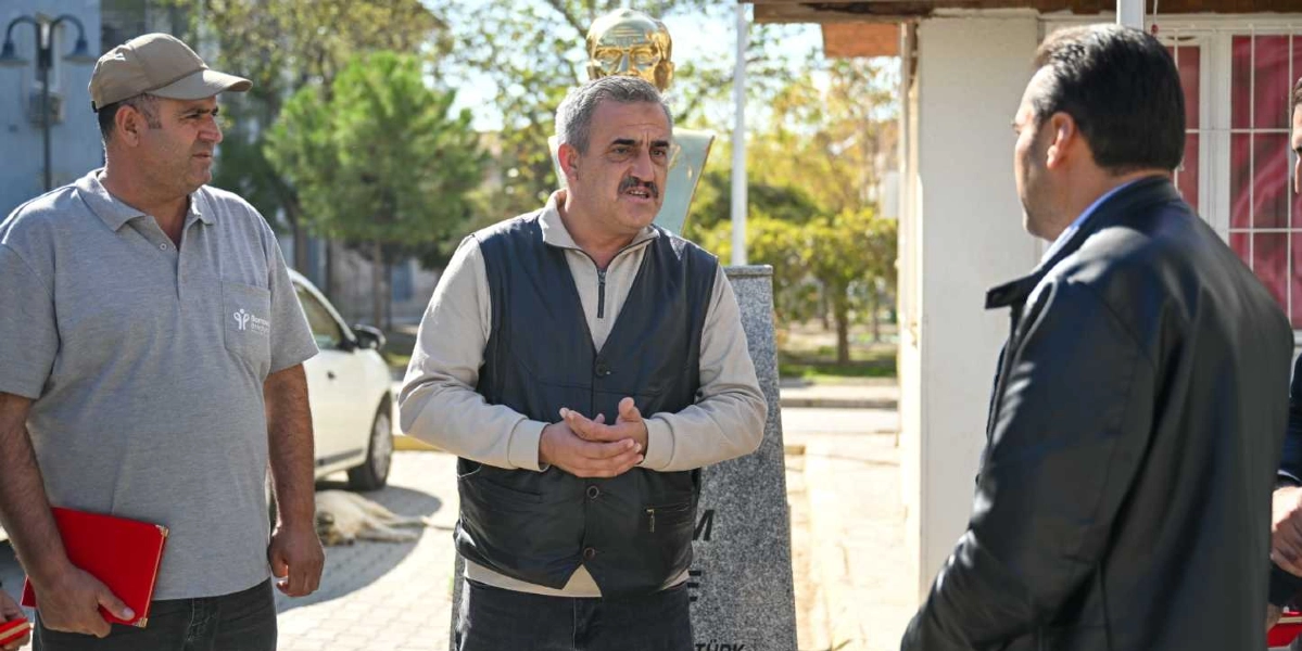 Açık havada, arkada Atatürk büstünün bulunduğu bir alanda üç erkek bir arada duruyor. Solda gri tişörtlü ve şapkalı bir kişi elinde kırmızı bir dosya tutarken, ortada bej renkli bir kazak ve siyah deri yelek giymiş bir adam konuşuyor. Sağ tarafta ise sırtı dönük, siyah deri ceketli bir başka kişi yer alıyor. Arka planda ağaçlar ve park edilmiş beyaz bir otomobil görülüyor.