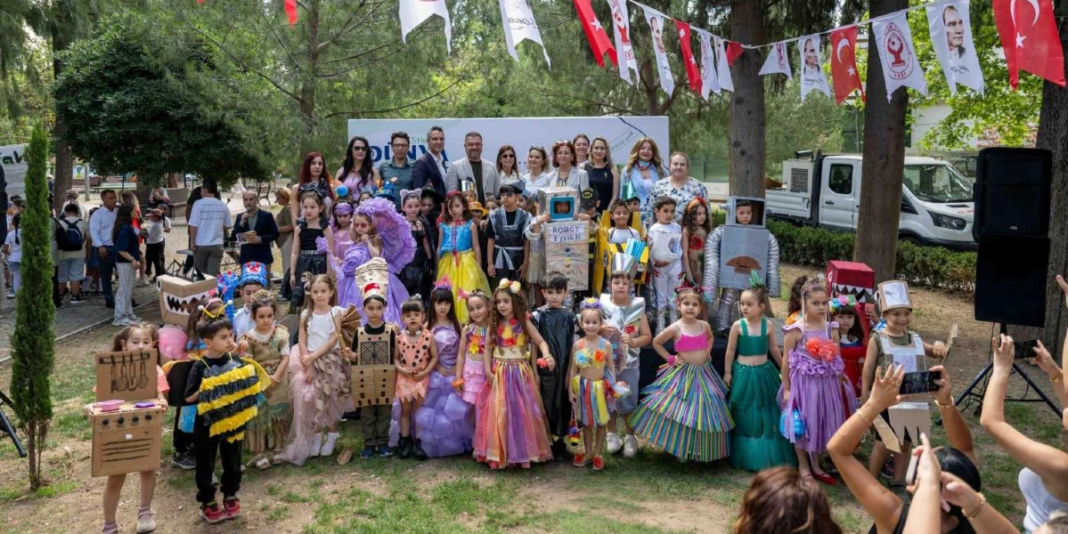 Açık havada düzenlenen bir etkinlikte, atık malzemelerden yapılmış geri dönüşüm kostümleri giyen çok sayıda çocuk ve arkalarında yetişkinler bir arada duruyor. Çocukların kostümleri arasında karton kutulardan yapılmış robotlar, mutfak gereçleri, renkli poşetlerden tasarlanmış elbiseler ve arı kostümü gibi yaratıcı kıyafetler dikkat çekiyor. Arka planda Bornova Belediyesi logolu bir pano, ağaçlar arasında asılı Türk bayrakları ve Atatürk görselleri bulunan flamalar yer alıyor. Ön planda bazı izleyiciler ellerindeki telefonlarla fotoğraf çekiyor.