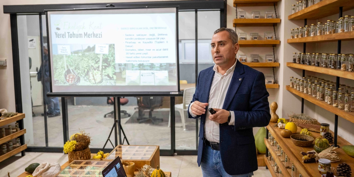 Koyu mavi ceketli bir erkek, Bornova Yerel Tohum Merkezi konulu bir sunum yaparken görülüyor. Arkasındaki projeksiyon perdesinde, Pınarbaşı ve Kayadibi'ndeki 6 dekarlık alanda 38 farklı yerel tohum çeşidinin üretilip paketlendiğini belirten bilgiler yer alıyor. Sunumun yapıldığı odanın duvarlarında, raflara dizilmiş çok sayıda küçük cam kavanoz içinde tohum örnekleri bulunuyor; ön plandaki masada ise çeşitli kabaklar, kuru bitkiler ve tohum sergileme kutuları yer alıyor.