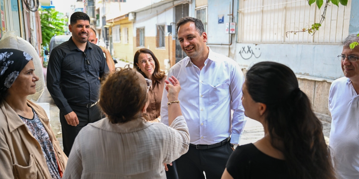 Çamdibi sokaklarında beyaz gömlekli bir erkek, çevresindeki mahalle sakinleriyle samimi bir şekilde sohbet ediyor. Grubun içinde gülümseyen kadınlar ve erkekler bulunurken, ön planda sırtı dönük bir kadın el hareketleriyle konuşuyor. Arka planda ise eski binaların olduğu dar bir sokak yer alıyor.