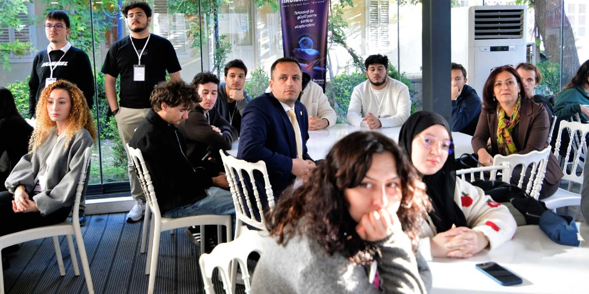 Aydınlık bir iç mekanda, beyaz masalar etrafında toplanmış gençlerden ve yetişkinlerden oluşan bir grup, dikkatle bir sunumu veya konuşmacıyı dinliyor. Katılımcıların bir kısmı masalarda otururken, bir kısmı ayakta duruyor. Arka planda üzerinde 