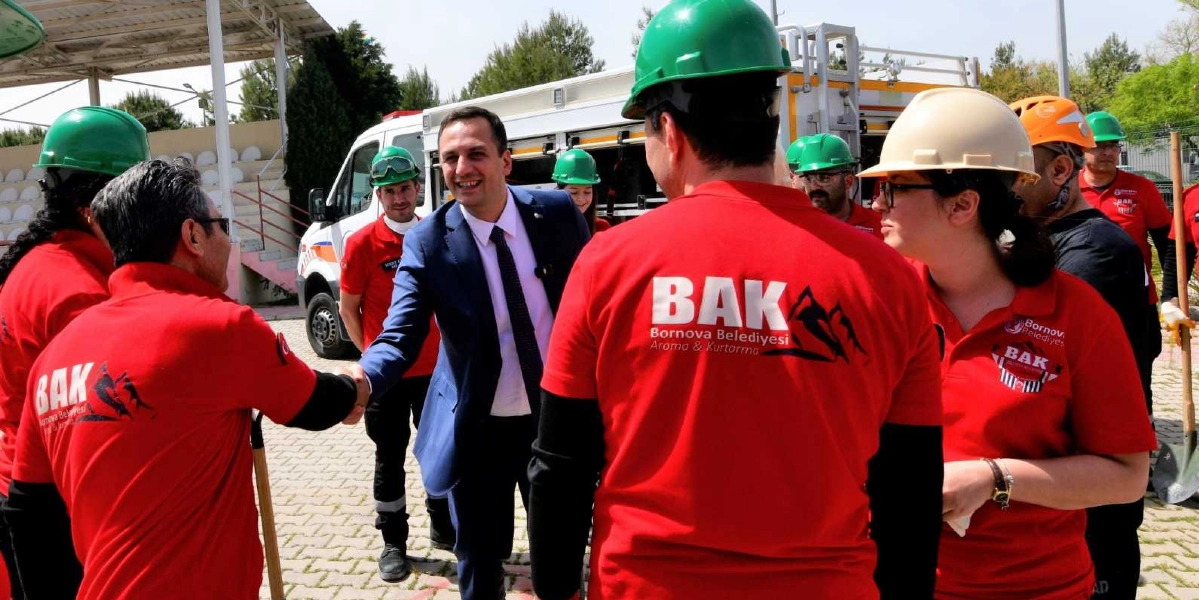 Mavi takım elbiseli bir yetkili, Bornova Belediyesi Arama ve Kurtarma (BAK) ekibinden kırmızı tişörtlü ve baretli bir personelle el sıkışıyor. Ekip üyelerinin üzerinde 