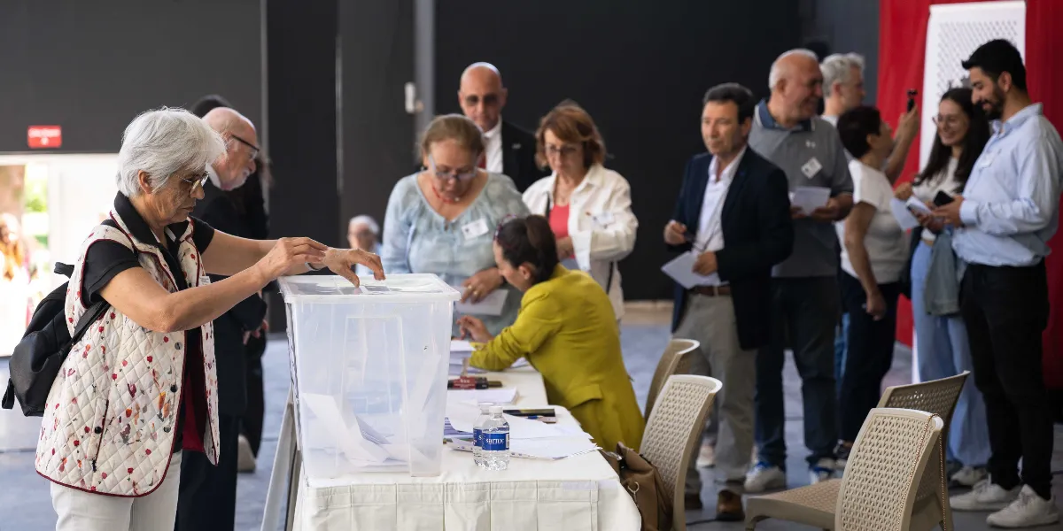 Atatürkçü Düşünce Derneği Genel Kurulu'nda, beyaz saçlı bir kadın şeffaf bir oy sandığına oy pusulasını atıyor. Sandığın başında sarı ceketli bir görevli otururken, arka planda sırasını bekleyen katılımcılar bulunuyor. Salonun genelinde resmi ve ciddi bir atmosfer hakim.