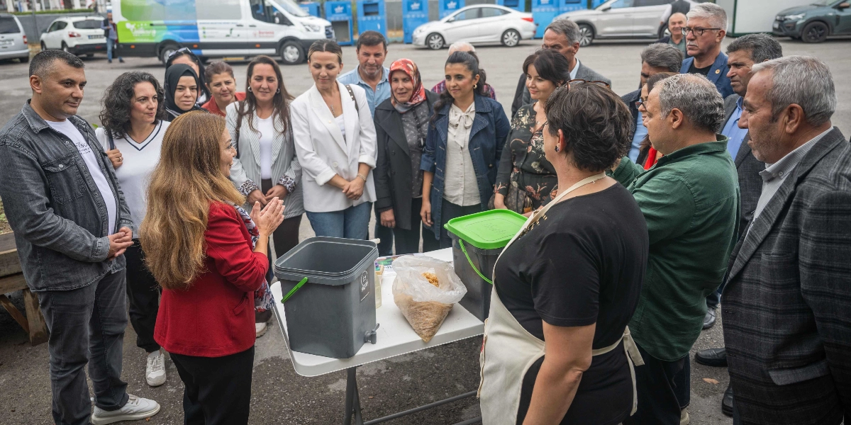 Açık havada, Bornova Belediyesi logolu bir aracın önünde toplanmış bir grup insan, sıfır atık uygulamaları üzerine yapılan bir bilgilendirme toplantısına katılıyor. Beyaz bir masanın üzerinde, içinde organik atıkların dönüştürülmesi için kullanılan musluklu gri bir kompost kovası ve bir poşet dolusu atık bulunuyor. Kırmızı ceketli bir kadın, gruptakilere bu ekipmanları ve süreci anlatırken, çevresindeki katılımcılar dikkatle onu dinliyor.