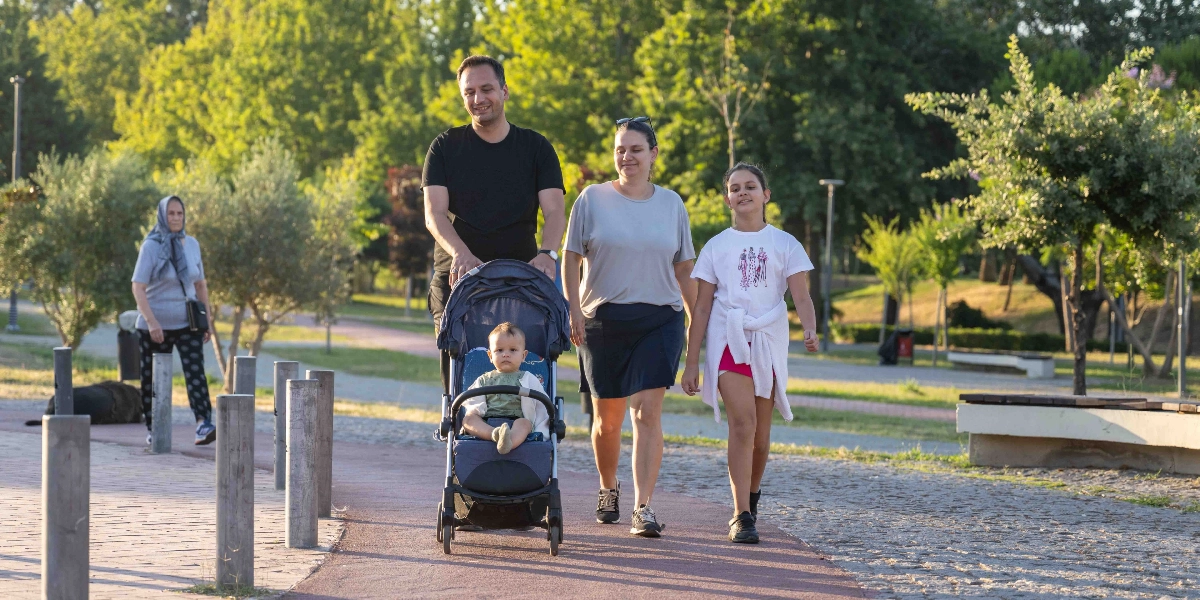 Yeşil bir parkta yürüyüş yapan bir aile; siyah tişörtlü bir erkek bebek arabasını sürerken, yanında gri tişörtlü bir kadın ve beyaz tişörtlü genç bir kız çocuğu yürüyor. Bebek arabasında küçük bir çocuk oturuyor. Arka planda ise parkta yürüyen başka bir kadın ve ağaçlar görülüyor.